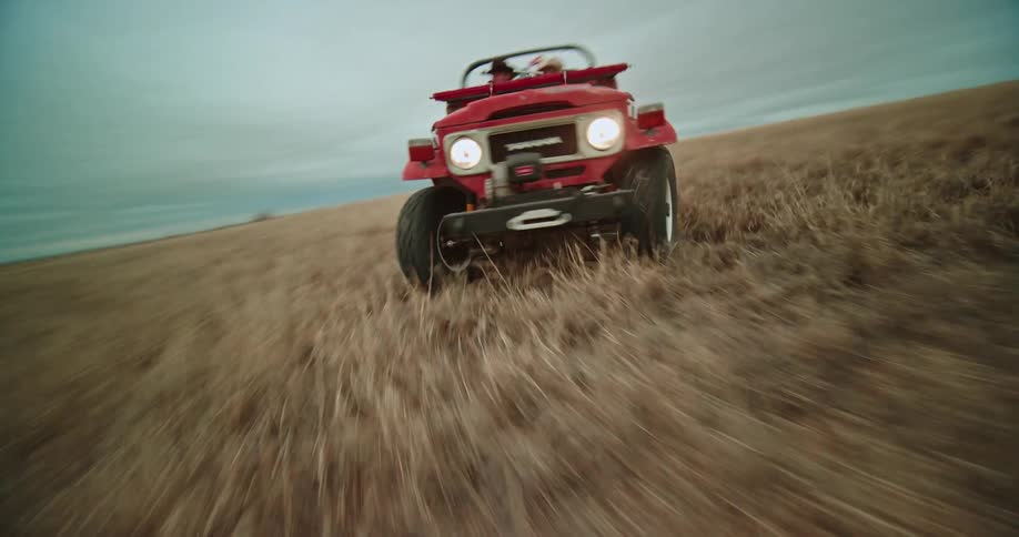 Two people sit in red jeep turning and speeding in field under overcast sky