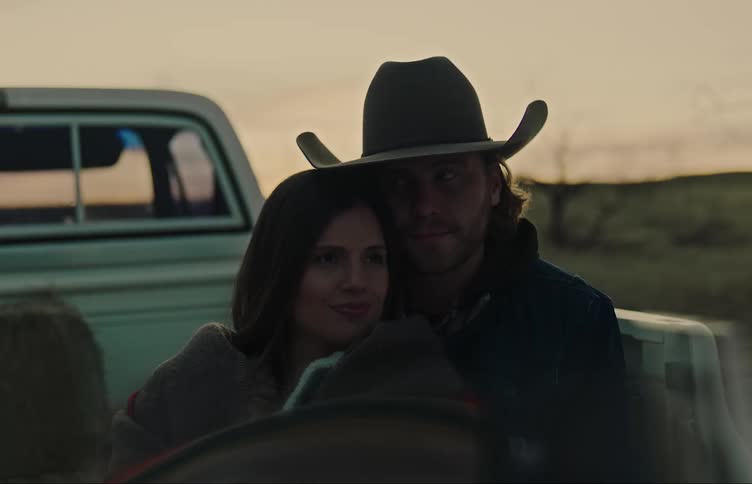 Man wearing cowboy hat cuddles with woman in back of pickup truck parked in field under a red tinted sky