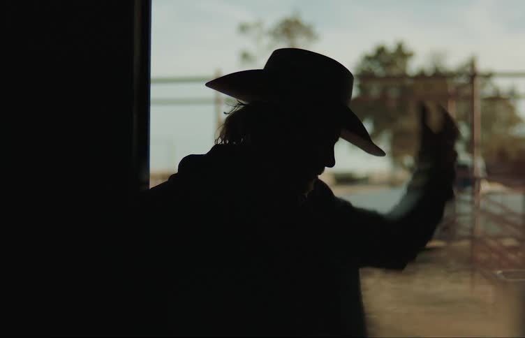 A silhouette of a man standing in a doorway outdoors putting his hat on and staring forward