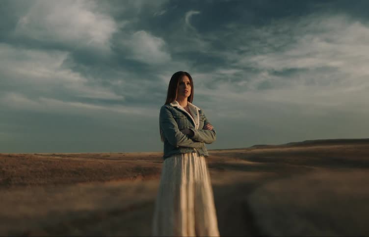 Woman with brown hair denim jacket and white dress stands arms crossed on dirt road in the field