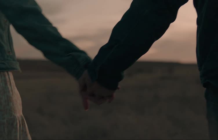 Two lovers are walking through a field at sunset on a cloudy evening and the woman smiles looking at her boyfriend while passing her left hand through her long hair