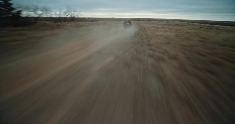 An openroofed vehicle speeds down a dirt road among a muted grassy plain in the wilderness