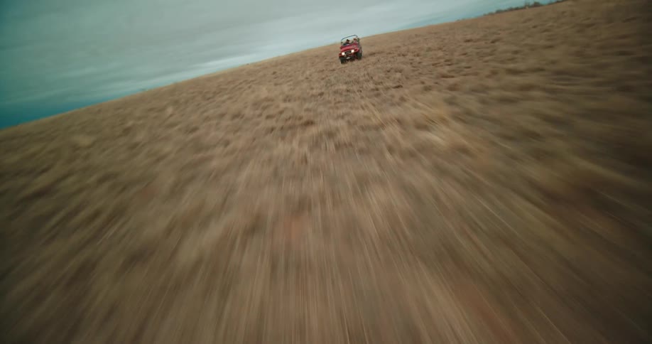 Two people drive across grassland in a red jeep under an overcast sky