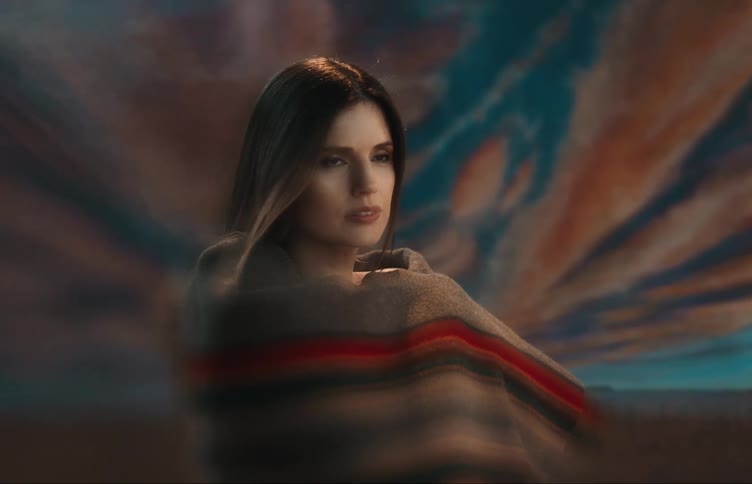 A woman is standing with a blanket around her shoulders as light shines on her face and her hair moves in the breeze