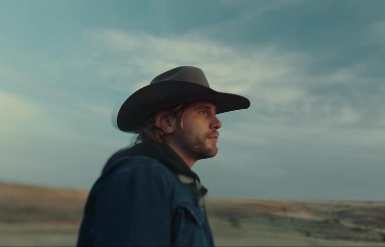 A man is standing in outside with his hand on his cowboy hat as he puts it on his head and looks up
