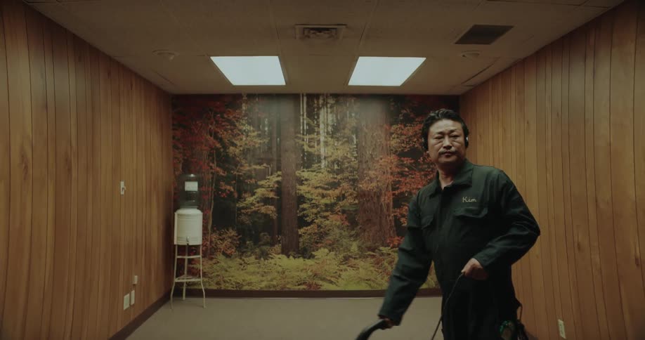 A man is wearing headphones as he vacuums a room with wood paneling and a forest picture on the wall