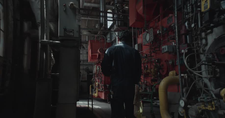 A man is walking in a hallway near machines as he pushes a mop bucket through the back room of a facility