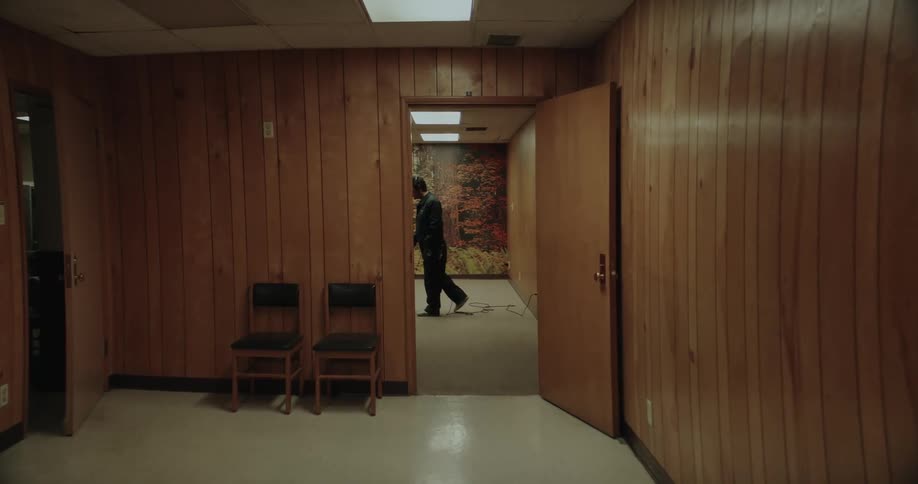 A man is wearing headphones as he vacuums a room with wood paneling and a forest picture on the wall