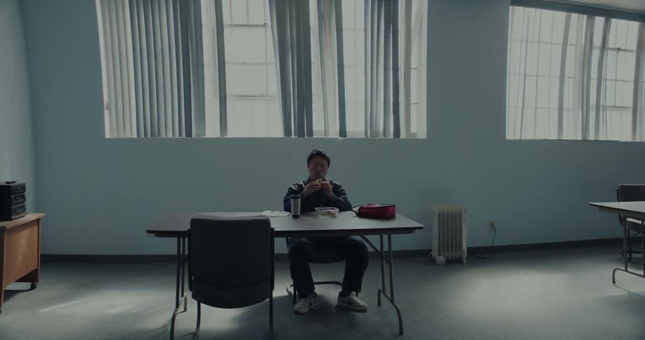 Someone walks in a hallway as a man is sitting at a table in a room eating his lunch as he holds his sandwich as drinks from his cup