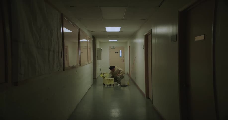 A man is standing next to a mop bucket as he mops a hallway and stops to look inside the bucket