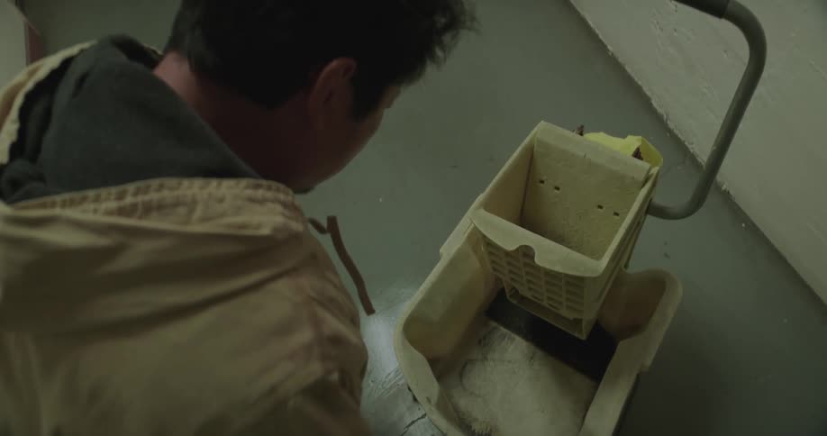 Man crouches near mop bucket in hallway and reaches for something in the bucket