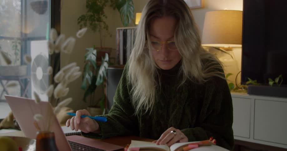 A woman works on a laptop while looking back and forth from the laptop to a well worn book