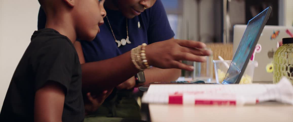 Mother with earrings and a nose ring and son with black t shirt sits at a table and uses a laptop in the kitchen