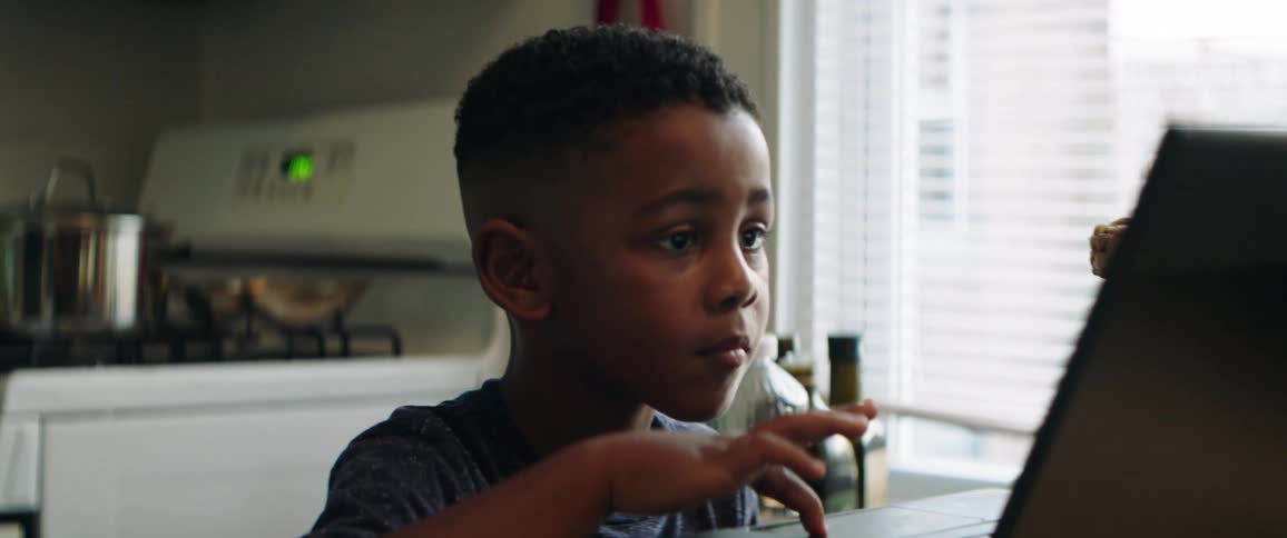 Young boy sitting at kitchen table looking at laptop screen while using the track pad on the keyboard