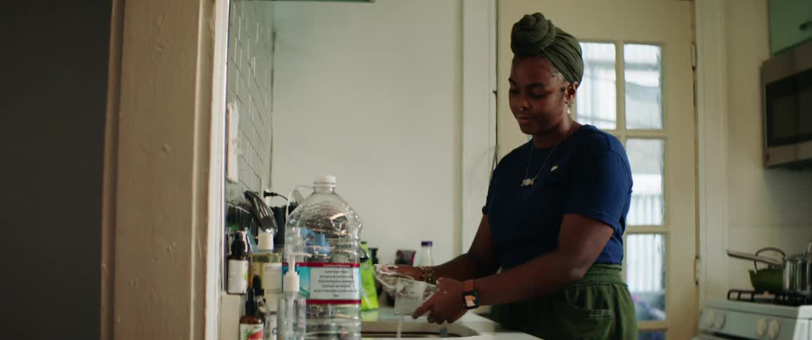 A woman in the kitchen washing a glass in the sink with a door and a stove in the background