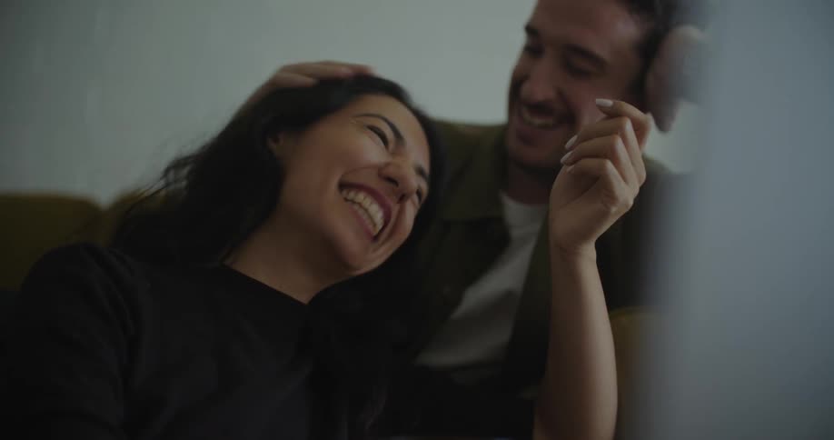 A man sits on a couch and strokes a womans hair as they talk and laugh and then he leans over and kisses her head