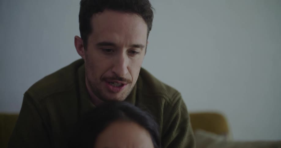 A woman is sitting on the floor in front of a table looking at a laptop computer as a man is sitting on the couch behind her looking over her head