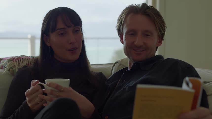 A man and woman sitting on a couch together he kisses her on the cheek while holding a book and she holds a mug