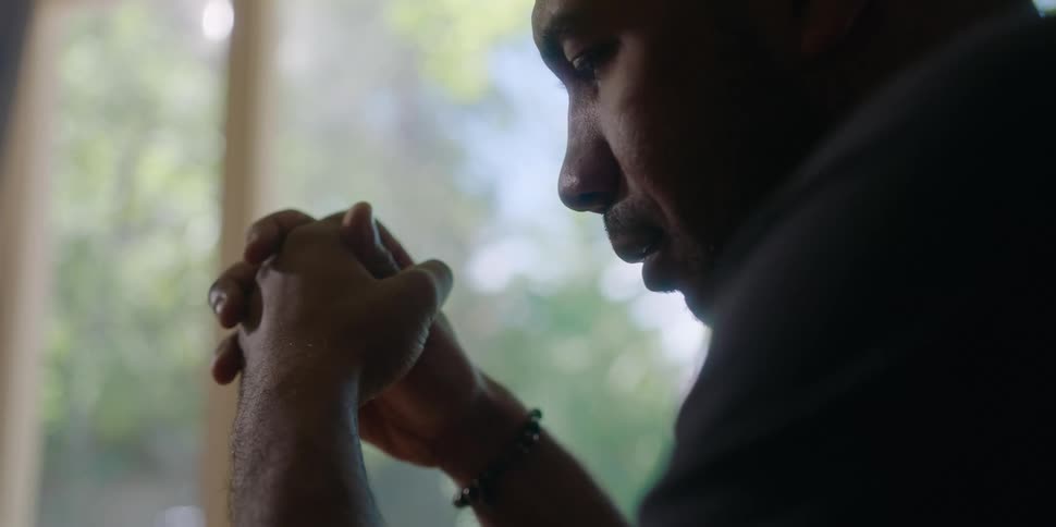 A man sits with his palms clasped next to a window