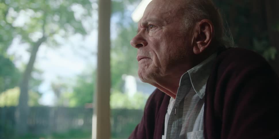 An elderly man sits in front of a large window at home and shakes the hand of someone across from him