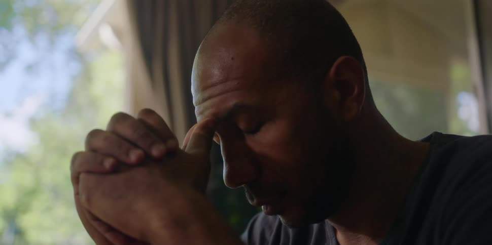 A man is sitting down with his hands clasped in front of him and he rests his forehead on his thumbs and thinks