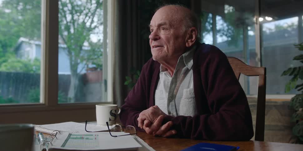 An elderly man sits at a table laughing and pointing as he talks