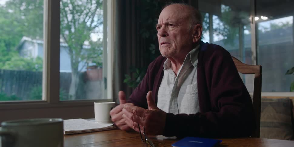 An elderly man at home shakes hands with a man across the table and then gets angry as he speaks