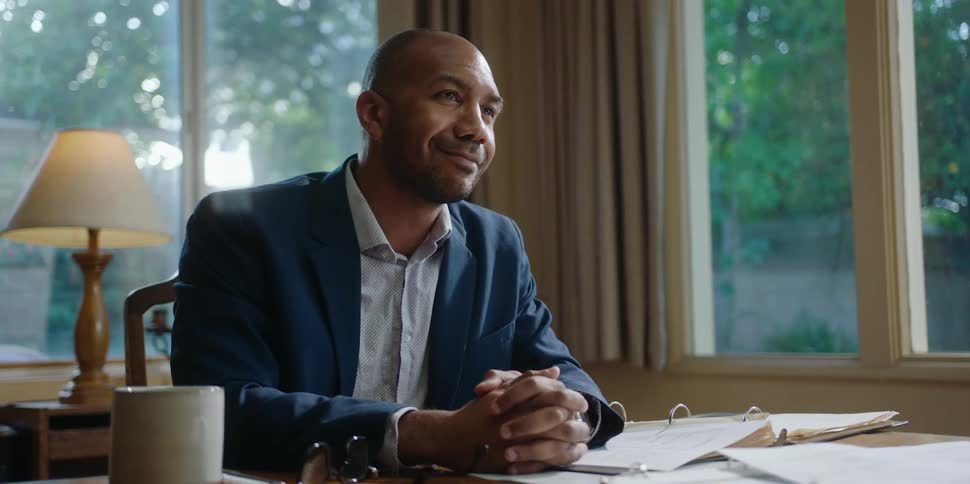 A man is sitting at a desk with an open notebook and smiling with his hands folded as the person across the desk looks at documents
