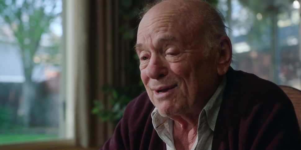An elderly man is sitting down moving his hands and smiling as he speaks
