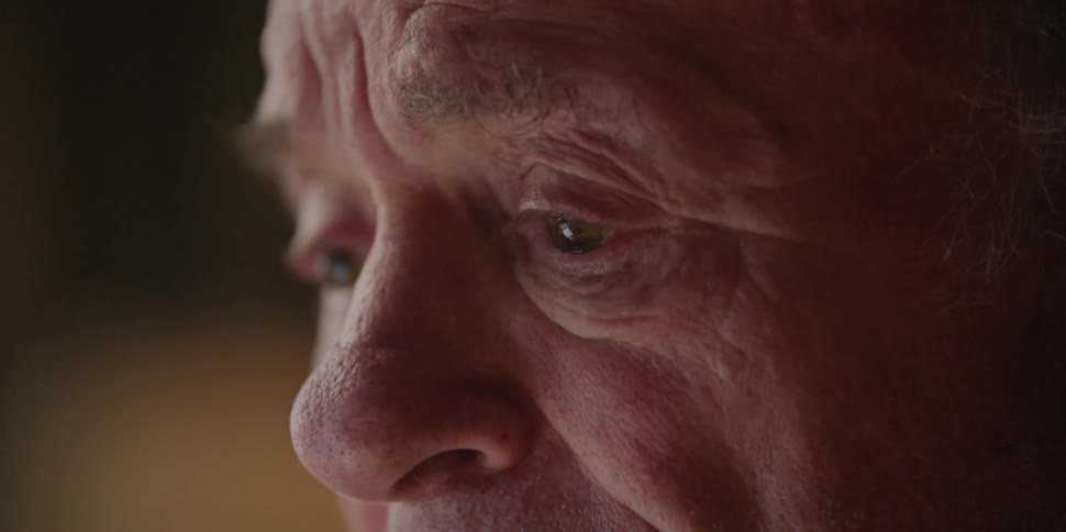 An elderly man blinks while talking to another person offcamera