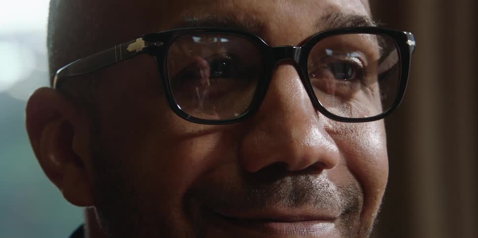 A man wearing glasses nods his head as another man speaks and that mans reflection is seen in the lenses