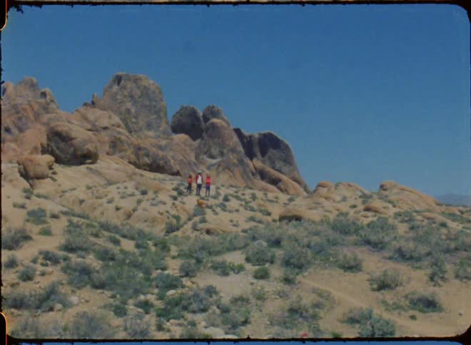 Three people are standing on a mountainside with their dog near huge boulders on the mountain under a clear blue sky