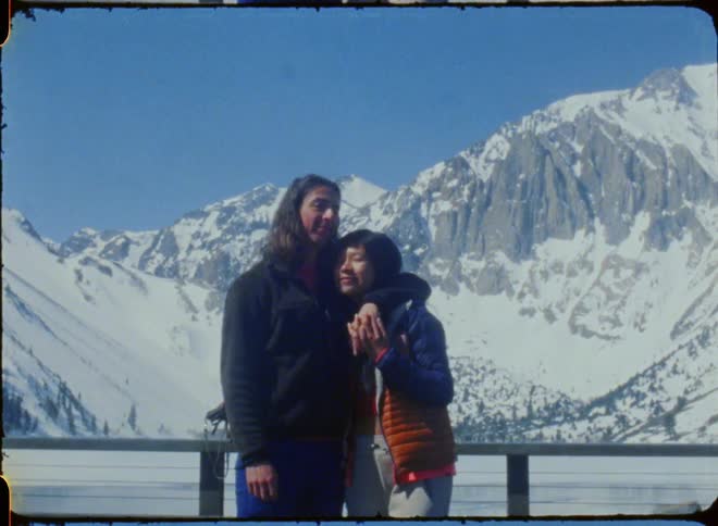 A man kisses his partner on the forehead while posing in front of a snow covered mountain
