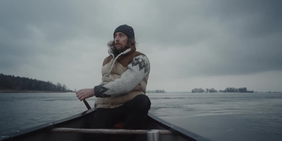 Man wearing quilted vest paddles canoe with forest covered shore in the distance on a gray overcast day