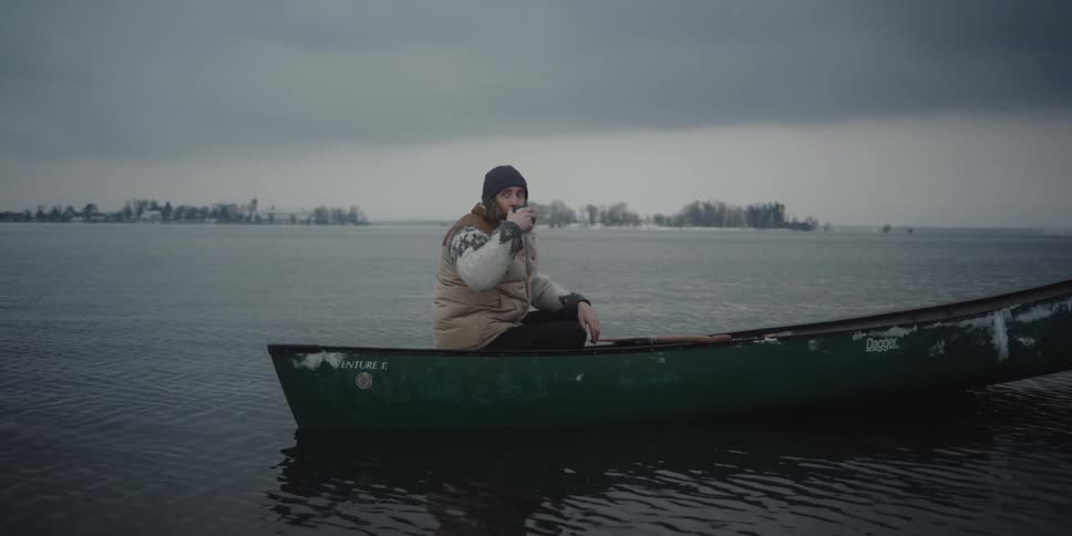 A pensive man wearing winter clothes sits in a small boat drinking from a mug