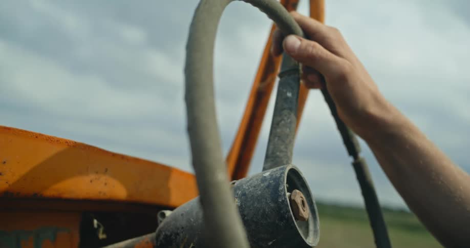 A man's hand holds a steering wheel of an old jeep as it travels in a rural area under a cloudy sky