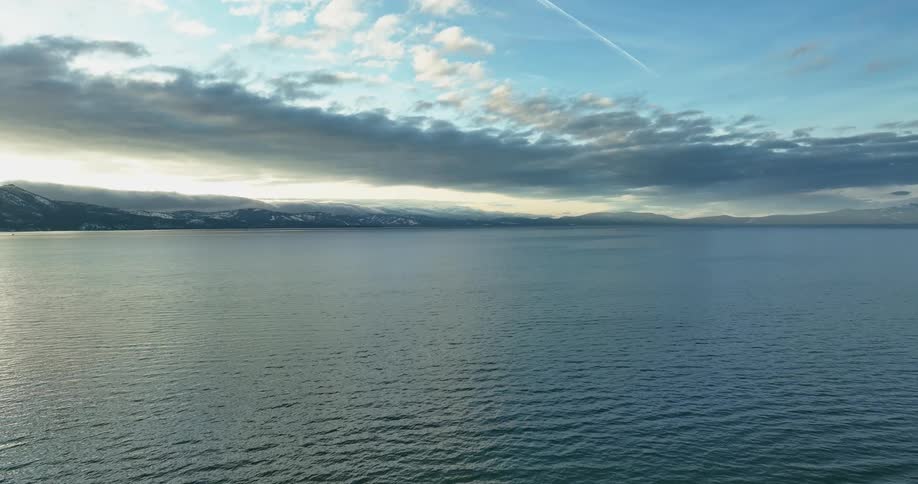 Light shines on the water as it ripples and waves and there are mountains in the distance under a cloudy sky