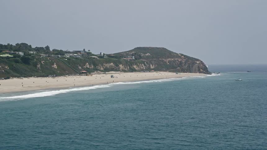 A low-flying 5K aerial footage captures the stunning coastline of Malibu California by Point Dume with a sandy beach underneath a cliff bustling with beachgoers and sunbathers on a hazy day