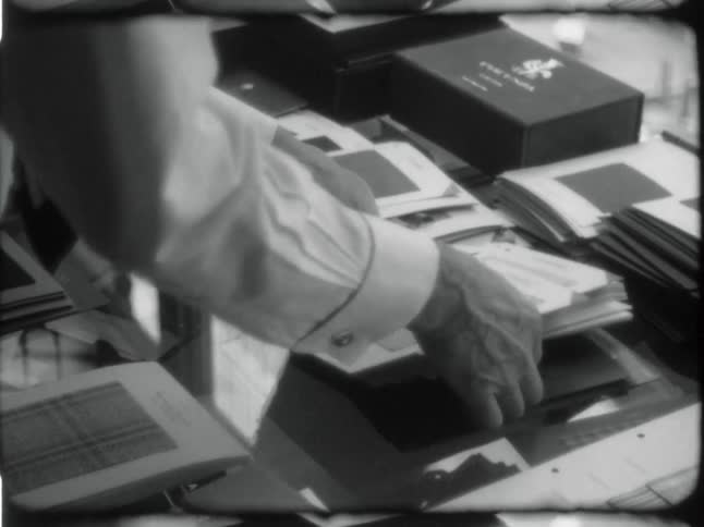 A black and white clip of an office worker wearing a dress shirt and tie while they arrange the notebooks on a glass office table