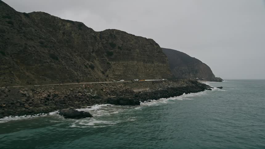 Aerial view of waves crashing into a scenic coastal road by Highway 1 in Malibu California with light traffic at daytime