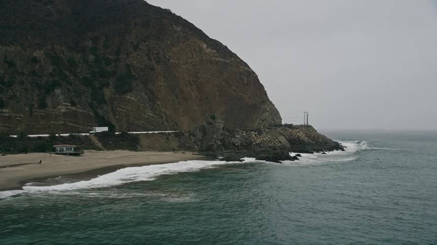 A low altitude aerial approach to waves crashing against rocks and the beach in Malibu California with a hazy atmosphere and rock formations