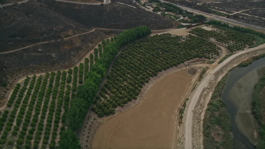 Aerial footage of a hazy day approaching orchards and farmland at the base of mountains near a country road in Point Mugu California