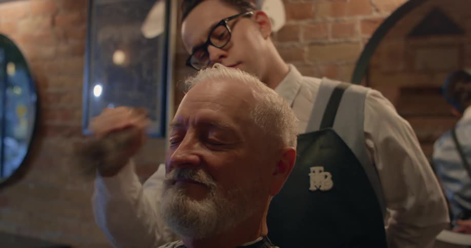 A man smiles with his eyes closed at a barbershop while the barber brushes loose hairs off his head