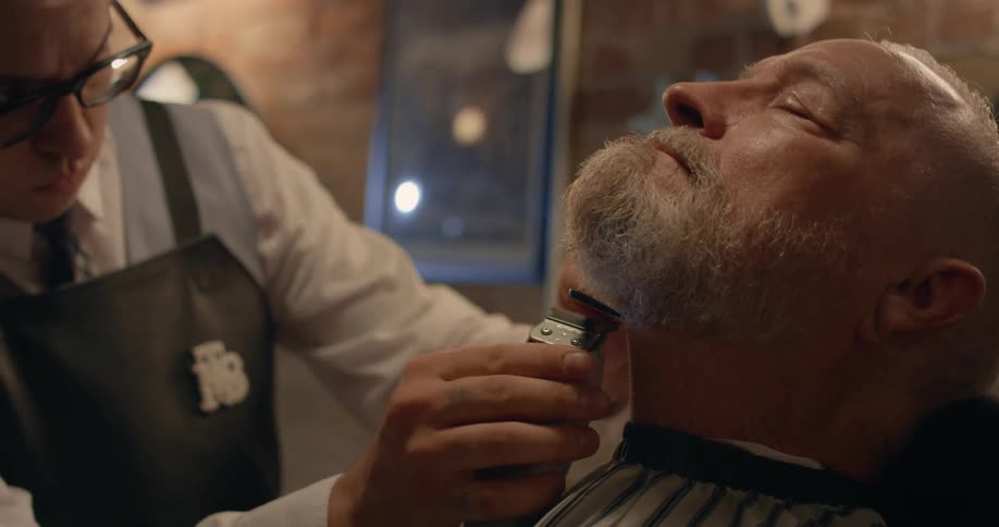 A man leans back as a barber trims his beard with electric clippers
