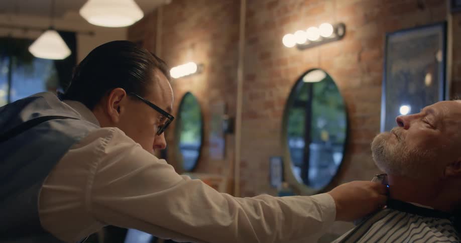 Man with glasses and white shirt and blue vest cuts mans beard with electric shaver in the barbershop