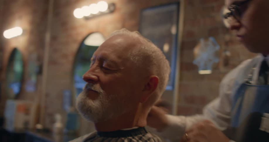 A man sits with his eyes closed at a barbershop smiling while the barber brushes the loose hairs off his head