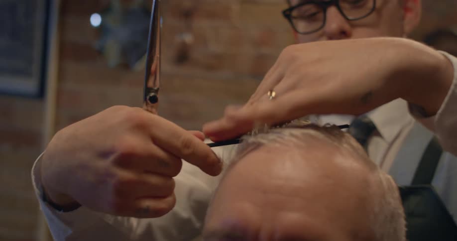 Barber with glasses and white shirt cuts mans gray hair with scissors and comb in the barbershop