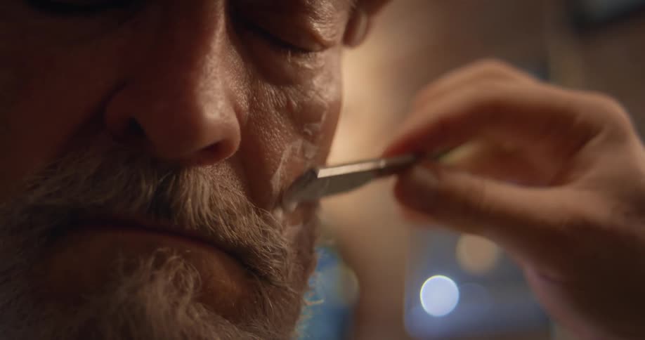 A bearded man closes his eyes as a barber carefully give him a shave
