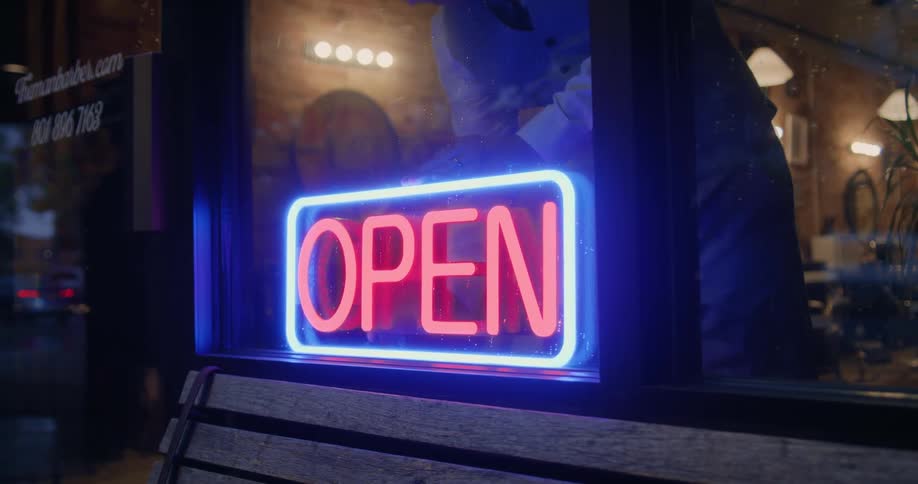A man inside a barbershop walks over to the neon sign in the window and turns it on as he opens for business