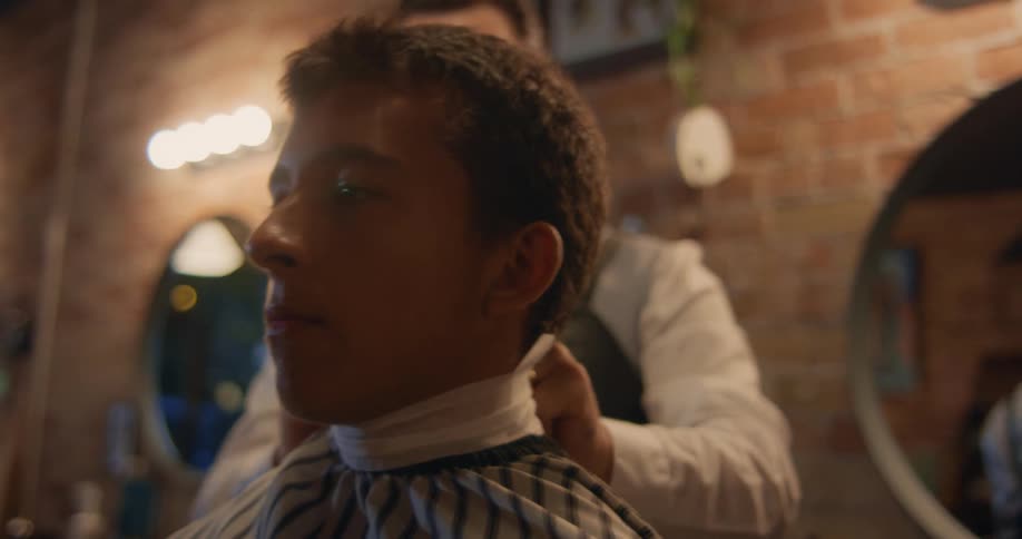 A man sitting in a barbers chair while the barber puts a towel around the customers neck and adjust the cape